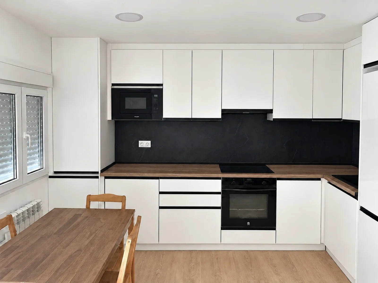 Cocina en L con muebles blancos sin tirador, frente negro y encimera de madera.